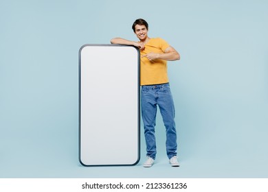 Full body young man wear yellow t-shirt point index finger on big mobile cell phone with blank screen workspace area isolated on plain pastel light blue background studio. People lifestyle concept. - Shutterstock ID 2123361236