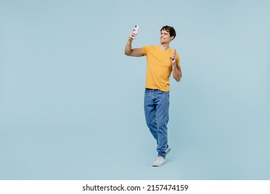 Full body young man wear yellow t-shirt doing selfie shot on mobile cell phone post photo on social network show v-sign isolated on plain pastel light blue background studio. People lifestyle concept