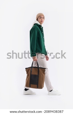 Full body young man wearing a white hoodie with green shirt with hat holding handbag walking in studio


