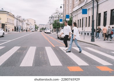 Full body of young man and woman in casual clothes holding hands while crossing street on crosswalk during date in city
