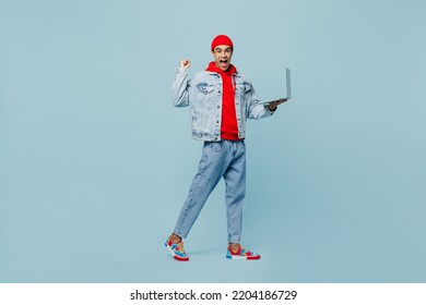 Full Body Young Middle Eastern IT Man He Wear Denim Jacket Red Hat Hold Use Work On Laptop Pc Computer Do Winner Gesture Isolated On Plain Pastel Light Blue Cyan Background People Lifestyle Concept