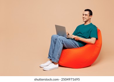 Full body young IT middle eastern man wear blue t-shirt casual clothes sit in bag chair hold use work on laptop pc computer isolated on plain pastel beige background studio portrait. Lifestyle concept - Powered by Shutterstock