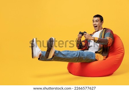 Full body young middle eastern man 20s he wear casual shirt white t-shirt sit in bag chair hold in hand play pc game with joystick console isolated on plain yellow background People lifestyle concept