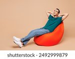 Full body young minded fun middle eastern man wear blue t-shirt casual clothes sit in bag chair hold hands behind head neck isolated on plain pastel beige background studio portrait. Lifestyle concept