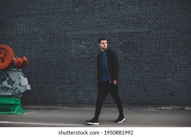 Full body of young modern hipster male in trendy outfit walking on street beside high dark brick wall and looking away