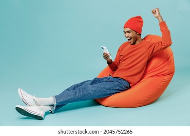 Full body young overjoye fun happy african american man wearing orange shirt hat sit in bag chair use hold mobile cell phone do winner gesture isolated on plain pastel light blue background studio.