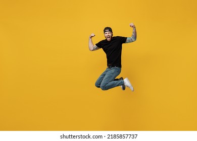Full body young overjoyed cool bearded tattooed man 20s he wears casual black t-shirt cap jump high do winer gesture isolated on plain yellow wall background studio portrait. People lifestyle concept