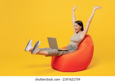 Full Body Young Overjoyed Happy Latin Woman 30s She Wear Basic Beige Tank Shirt Sit In Bag Chair Hold Use Work On Laptop Pc Computer Spread Raise Up Hands Isolated On Plain Yellow Backround Studio.