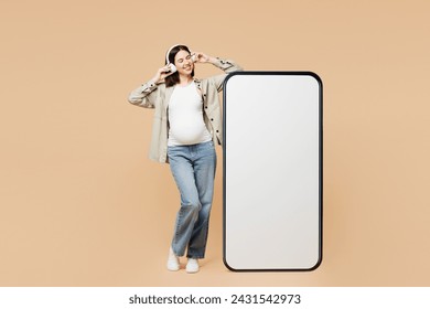 Full body young pregnant woman future mom wear grey shirt headphones belly stomach tummy with baby big blank screen mobile cell phone listen music isolated on plain beige background. Maternity concept - Powered by Shutterstock