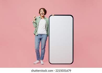 Full body young proactive woman wear green shirt white t-shirt near big huge blank screen mobile cell phone smartphone with mockup area point finger up isolated on plain pastel light pink background - Shutterstock ID 2215031679