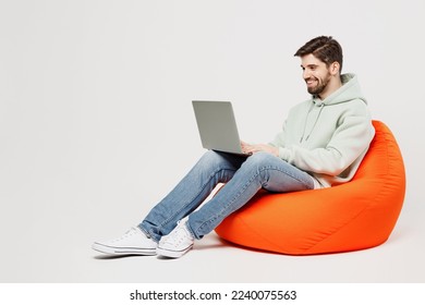 Full body young programmer happy caucasian IT man wear mint hoody sit in bag chair hold use work on laptop pc computer isolated on plain solid white background studio portrait People lifestyle concept