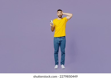 Full body young sad shocked fearful caucasian man wear yellow t-shirt use mobile cell phone hold head look camera isolated on plain pastel light purple background studio portrait. Lifestyle concept