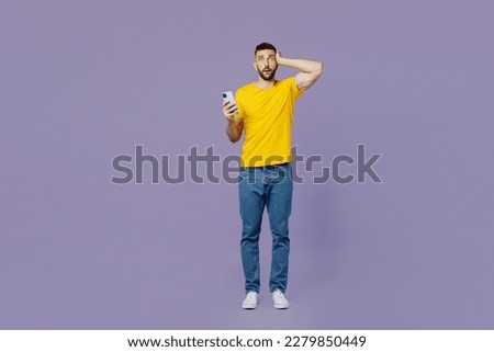 Full body young sad shocked fearful caucasian man wear yellow t-shirt use mobile cell phone hold head look camera isolated on plain pastel light purple background studio portrait. Lifestyle concept