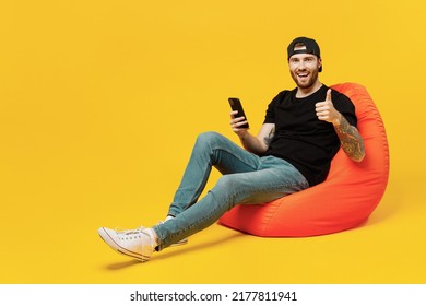 Full body young satisfied bearded tattooed man 20s he wears casual black t-shirt cap sit in bag chair hold in hand use mobile cell phone show thumb up isolated on plain yellow wall background studio