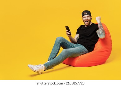 Full body young satisfied bearded tattooed man he wears casual black t-shirt cap sit in bag chair hold in hand use mobile cell phone do winner gesture isolated on plain yellow wall background studio