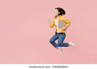 Full body young satisfied cheerful cool IT woman of Asian ethnicity wear yellow shirt white t-shirt jump high hold use work on laptop pc computer isolated on plain pastel light pink background studio