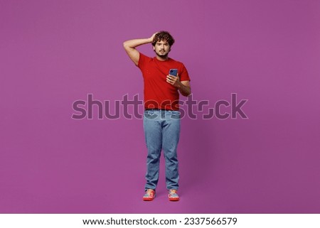 Full body young shocked frightened unhappy Indian man wear red t-shirt casual clothes hold head use mobile cell phone look camera isolated on plain purple background studio portrait. Lifestyle concept