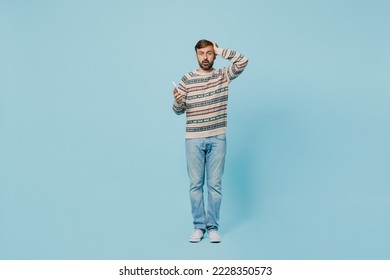 Full body young shocked sad caucasian man 30s he wear sweater hold in hand use mobile cell phone hold head isolated on plain pastel light blue cyan background studio portrait. People lifestyle concept