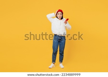 Full body young shocked sad woman of Asian ethnicity wear white padded windbreaker jacket red hat hold head use mobile cell phone isolated on plain yellow background studio. People lifestyle concept