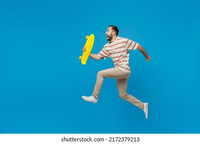 Full body young side view happy fun cool caucasian man 20s wear orange striped t-shirt hold yellow skateboard jump high run isolated on plain blue background studio portrait. People lifestyle concept