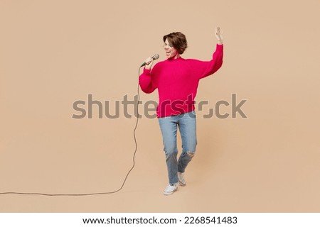 Full body young singer expressive woman wear pink sweater sing song in microphone at karaoke club raise up hand isolated on plain pastel light beige background studio portrait People lifestyle concept