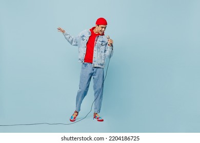 Full body young singer middle eastern man 20s he wear denim jacket red hat sing song in microphone at karaoke club isolated on plain pastel light blue cyan background studio. People lifestyle concept