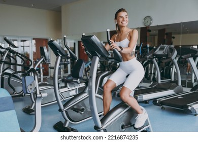 Full body young skinny strong sporty athletic sportswoman caucasian woman in white sportswear earphones listen music look aside warm up train use exercise bike in gym indoors. Workout sport concept - Powered by Shutterstock