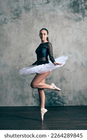 Full body of young slim ballerina in white tutu and pointe standing on tiptoes against shabby wall looking at camera