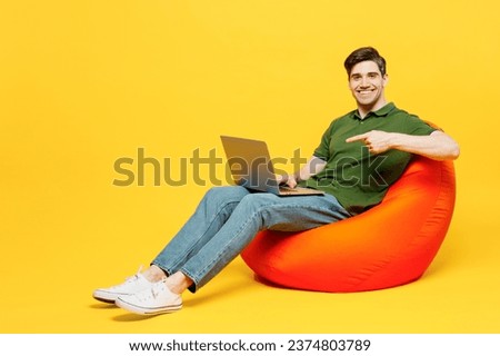 Full body young smart programmer happy IT man wear green t-shirt casual clothes sit in bag chair hold use work point finger on laptop pc computer isolated on plain yellow background. Lifestyle concept