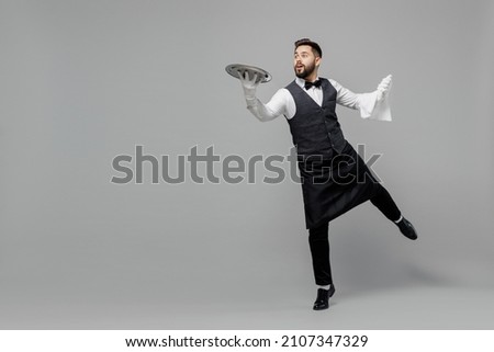 Full body young smiling barista male waiter butler man wear white shirt vest elegant uniform work at cafe carrying walk give metal tray isolated on plain grey background Restaurant employee concept