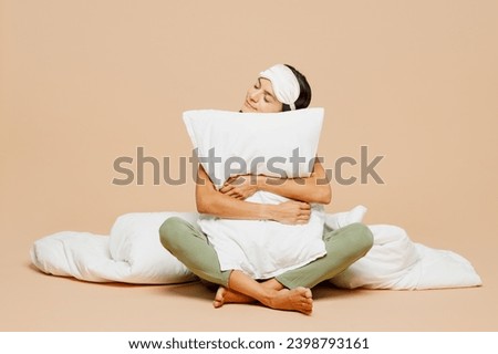 Full body young smiling calm Latin woman wears pyjamas jam sleep eye mask rest relax at home sitting with pillow hold duvet close eyes isolated on plain beige background. Good mood night nap concept