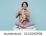 Full body young smiling cheerful happy woman 20s in brown shirt sit on white chair use point finger on mobile cell phone isolated on pastel plain light blue background studio People lifestyle concept