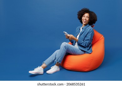 Full Body Young Smiling Cheerful Student Fun Happy Black Woman In Casual Clothes Shirt White T-shirt Sit In Bag Chair Hold Use Mobile Cell Phone Isolated On Plain Dark Blue Background Studio Portrait.