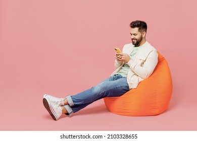 Full body young smiling cheerful happy man 20s in trendy jacket shirt sit in bag chair hold in hand use mobile cell phone isolated on plain pastel light pink background studio People lifestyle concept - Shutterstock ID 2103328655