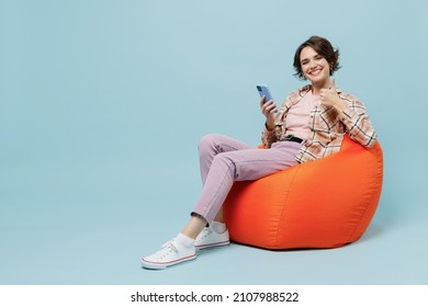 Full Body Young Smiling Cheerful Fun Happy Woman 20s In Brown Shirt Sit In Bag Chair Hold Use Mobile Cell Phone Show Thumb Up Gesture Isolated On Pastel Plain Light Blue Background Studio Portrait.