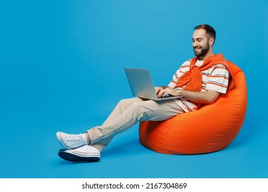 Full Body Young Smiling Cheerful Fun Man 20s In Orange Striped T-shirt Sit In Bag Chair Hold Use Work On Laptop Pc Computer Isolated On Plain Blue Background Studio Portrait. People Lifestyle Concept