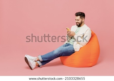 Full body young smiling cheerful happy man 20s in trendy jacket shirt sit in bag chair hold in hand use mobile cell phone isolated on plain pastel light pink background studio People lifestyle concept