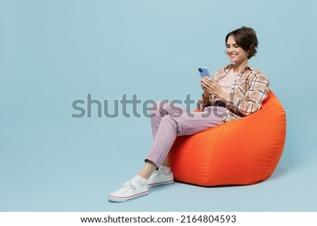 Full body young smiling cheerful happy woman 20s in brown shirt sit in bag chair hold use mobile cell phone isolated on pastel plain light blue background studio portrait. People lifestyle concept.