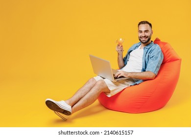 Full Body Young Smiling Cool Happy Man In Blue Shirt White T-shirt Sit In Bag Chair Hold Use Work On Laptop Pc Computer Alcohol Cocktail Martini Isolated On Plain Yellow Background Studio Portrait.