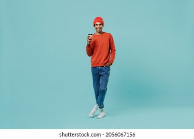 Full body young smiling happy african american man 20s wear orange shirt hat hold takeaway delivery craft paper brown cup coffee to go isolated on plain pastel light blue background studio portrait.