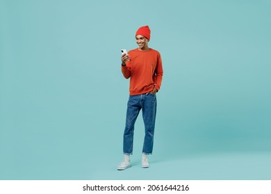 Full body young smiling happy african american man 20s in orange shirt hat use hold mobile cell phone chatting isolated on plain pastel light blue background studio portrait. People lifestyle concept