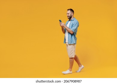 Full body young smiling happy caucasian man 20s wearing blue shirt white t-shirt hold in hand use mobile cell phone walk isolated on plain yellow background studio portrait. People lifestyle concept