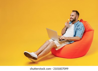 Full Body Young Smiling Happy Man In Blue Shirt White T-shirt Sit In Bag Chair Hold Use Work On Laptop Pc Computer Drink Alcohol Cocktail Martini Isolated On Plain Yellow Background Studio Portrait.