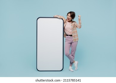 Full body young smiling happy woman 20s in casual brown shirt stand near big mobile cell phone with blank screen workspace area do winner gesture isolated on pastel plain light blue color background. - Shutterstock ID 2115785141
