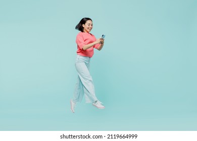 Full Body Young Smiling Happy Woman Of Asian Ethnicity 20s Wearing Pink Sweater Jump High Hold In Hand Mobile Cell Phone Isolated On Pastel Plain Light Blue Color Background. People Lifestyle Concept