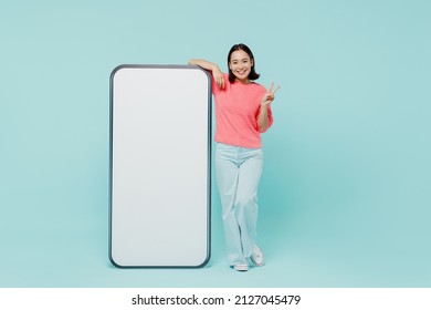Full body young smiling happy woman of Asian ethnicity 20s in pink sweater stand near big mobile cell phone with blank screen workspace area show v-sign isolated on pastel plain light blue background