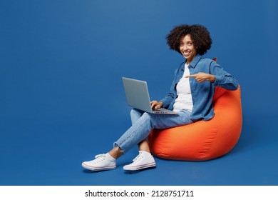 Full Body Young Smiling Happy Black Woman In Casual Clothes Shirt White T-shirt Sit In Bag Chair Hold Use Work Poing Finger On Laptop Pc Computer Isolated On Plain Dark Blue Background Studio Portrait