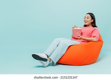 Full Body Young Smiling Happy Woman Of Asian Ethnicity 20s Wear Pink Sweater Sit In Bag Chair Hold Popcorn Bucket Watch Movie Film At Home Isolated On Pastel Plain Light Blue Color Background Studio