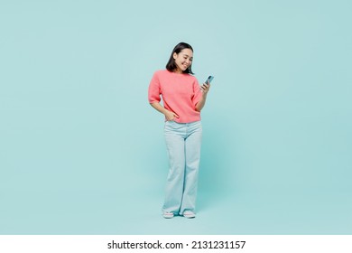 Full body young smiling happy woman of Asian ethnicity 20s wearing pink sweater hold in hand use mobile cell phone isolated on pastel plain light blue color background studio People lifestyle concept
