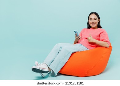 Full body young smiling happy woman of Asian ethnicity 20s wearing pink sweater sit in bag chair hold in hand use mobile cell phone show thumb up gesture isolated on pastel plain light blue background